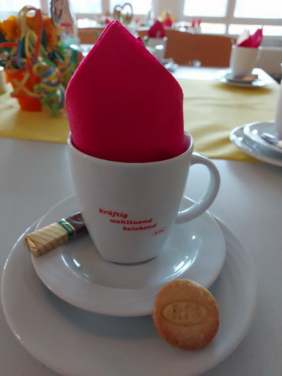 Tasse mit roter Serviette, Keks und Praline auf weißem Teller vor gedecktem Tisch.
