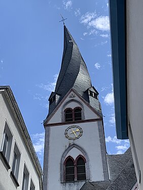 Kirchturm mit verdrehtem, spitz zulaufendem Dach vor blauem Himmel zwischen zwei Gebäuden