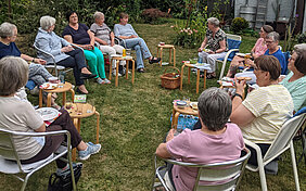 Eine Gruppe von Frauen sitzt auf Stühlen im Kreis an einem Sommertag in einem Garten, neben sich jeweils kleine Tische und darauf Trinkgläser und Getränkeflaschen.