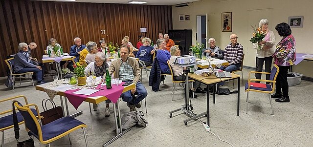 Mehrere ältere Personen sitzen an Tischen mit Tischdecken und Blumen in einem Veranstaltungsraum, zwei Frauen stehen vorne, eine hält einen Blumenstrauß, ein Beamer steht auf einem Rollwagen in der Mitte des Raums.