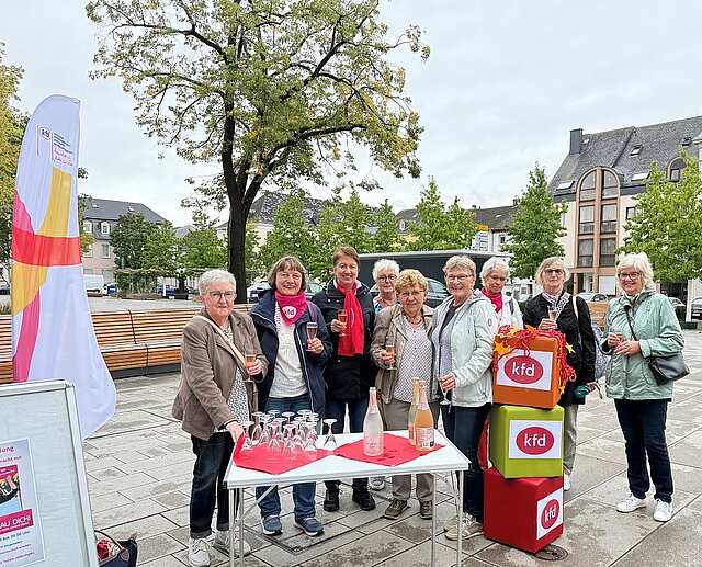  Eine Gruppe von Frauen, die in die Kamera schauen und Gläser in den Händen halten, vor Ihnen ein Tisch mit weiteren Gläsern und Getränkeflaschen. Sie stehen auf einem großen Platz, links der Gruppe ist eine Beachflagg, rechts vor der Gruppe stehen drei übereinander gestapelte Würfel, auf deren Seiten "kfd" geschrieben ist.