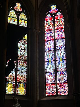 Zwei hohe gotische Buntglasfenster mit symmetrischen, farbigen Mustern in einer dunklen Kirchenwand