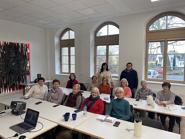 Gruppe von Menschen unterschiedlichen Alters sitzt an zusammengestellten Tischen in einem hellen Raum mit großen Fenstern und einem Laptop mit Beamer auf dem Tisch