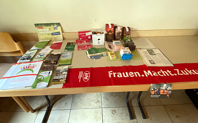Auf einem Tisch liegen verschiedene Faltblätter mit Informationen zu Fairem Handel und fair gehandelter Schokolade. Außerdem mehrere Schokoladentafeln verschiedener Sorten. Im Vordergrund an der Tischkante liegt ein Banner aus rotem Stoff, auf dem in weißer Schrift kfd - Katholische Frauengemeinschaft Deutschlands und Frauen.Macht.Zukunft geschrieben ist.