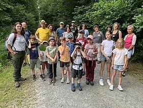 Gruppe von Erwachsenen und Kindern steht auf einem Waldweg, umgeben von dichtem Grün