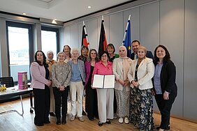 Gruppe von Menschen in einem Raum mit drei Fahnen im Hintergrund, eine Person hält eine Urkunde.