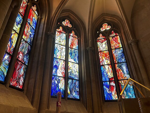 Mehrere hohe, bunte Glasfenster mit abstrakten und figürlichen Motiven in einer Kirche