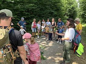 Gruppe von Erwachsenen und Kindern steht auf einem Waldweg, eine Frau mit rotem Rucksack hält Blätter in der Hand und spricht