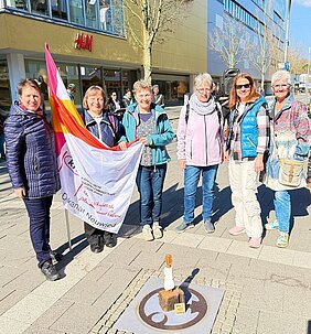 Sechs Personen stehen auf einem Bürgersteig vor einem Geschäft, eine Person hält eine Fahne mit dem Schriftzug 'Dekanat Neuwied', vor ihnen steht eine kleine Skulptur.