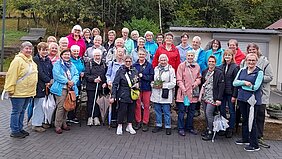 Eine Gruppe von Frauen in bunter Outdoorbekleidung steht auf gepflastertem Platz vor Büschen und einem kleinen Gebäude