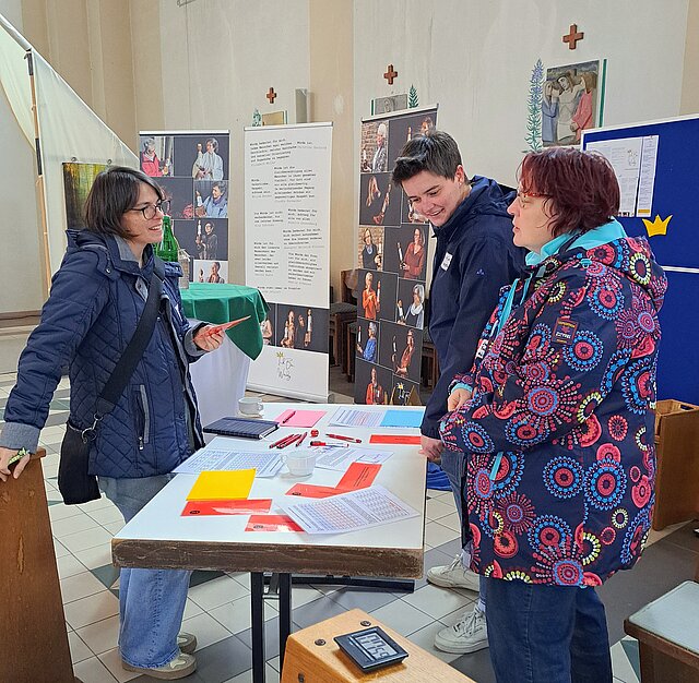 3 Frauen stehen um einen Tisch mit Spielmaterial und diskutieren. Im Hintergrund ist eine Ausstellung zu sehen.