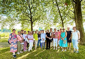 Gruppe von Frauen unterschiedlichen Alters steht im Schatten großer Bäume auf einer Wiese an einem sonnigen Tag