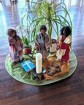 Aufnahme einer gestalteten Kreismitte bestehend aus drei Puppen im Hlabkreis stehend, einer weißen Kerze in einem Kerzenständer, einer Bibel, einem christlichen Holzkreuz und Blumen in weißen Blumentöpfen. Aufnahme einer gestalteten Kreismitte bestehend aus drei Puppen im Hlabkreis stehend, einer weißen Kerze in einem Kerzenständer, einer Bibel, einem christlichen Holzkreuz und Blumen in weißen Blumentöpfen.