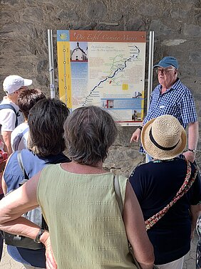 Gruppe von Menschen vor einer Informationstafel mit Karte und Text an einer Steinwand, ein Mann mit blauer Kappe erklärt etwas