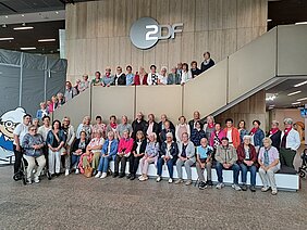 Eine Reisegruppe aus Männern und Frauen, teils sitzend, teils stehend, im Foyer des ZDF. Im Hintergrund ist an einer Wand das Logo des ZDF angebracht Eine Reisegruppe aus Männern und Frauen, teils sitzend, teils stehend, im Foyer des ZDF. Im Hintergrund ist an einer Wand das Logo des ZDF angebracht