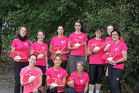 Gruppe von zehn Frauen in pinkfarbenen T-Shirts mit Fächern vor grünem Buschwerk