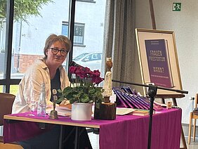 Eine Frau sitzt an einem Tisch auf dem mehrere Bücher liegen und der mit einer lila Tischdecke, einer kleinen Skulptur und Blumen in einem weißen Übertopf dekoriert ist. Sie spricht in ein Mikrofon. Auf einer Halterung rechts neben dem Tisch steht ein Plakat, das den vorderen Einband eines Buchs zeigt, Titel "Frauenpriestertum - wann?".