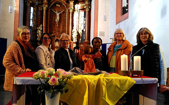 Sechs Frauen stehen hinter einem mit gelbem Tuch und Blumen geschmückten Altar in einer Kirche mit goldenem Altar im Hintergrund