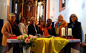 Sechs Frauen stehen hinter einem mit gelbem Tuch und Blumen geschmückten Altar in einer Kirche mit goldenem Altar im Hintergrund
