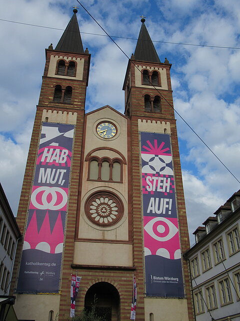 Kirche mit zwei hohen Türmen, an denen große Banner mit den Worten 'HAB MUT' und 'STEH AUF!' hängen