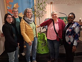 Fünf Frauen stehen in einer Kirche um einen jungen Baum mit grünem Schutzsack und lächeln in die Kamera