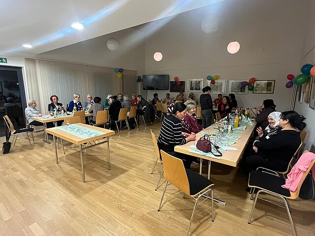 Mehrere Personen sitzen an zwei langen Tischen in einem hellen Raum mit Holzboden und weißen Wänden, an denen bunte Luftballons hängen.