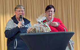 Zwei Frauen stehen an einem Stehtisch und schauen Richtung Betrachter:in. Eine von Ihnen hält ein Mikrofon in ihrer rechten Hand und spricht hinein. Auf dem Stehtisch im Vordergrund liegt ein verpacktes Geschenk.