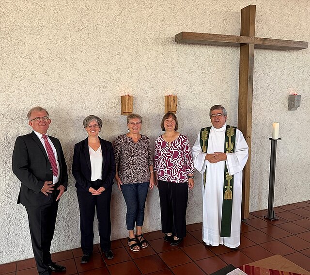 Fünf Personen stehen nebeneinander vor einer hellen Wand mit einem großen Holzkreuz, darunter ein Mann im schwarzen Anzug, drei Frauen in lässiger Kleidung und ein Geistlicher in weißem Gewand mit grünem Stola.