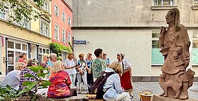 Gruppe von Menschen sitzt und steht in einem Innenhof neben einer großen Steinskulptur einer Frau mit einer Katze