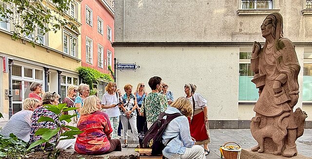 Gruppe von Menschen sitzt und steht in einem Innenhof neben einer großen Steinskulptur einer Frau mit einer Katze