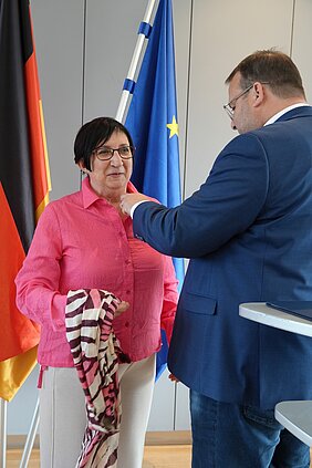 Zwei Personen bei einer Auszeichnung vor den Flaggen Deutschlands und der Europäischen Union.