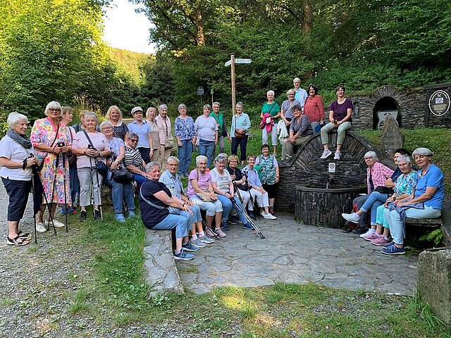 Eine Gruppe von Frauen sitzen bei sonnigem Wetter im Halbkreis um eine Quelle. Ein Hinweisschild im Hintegrund rechts trägt die Aufschrift "Venus-Charlotten Brunnen".