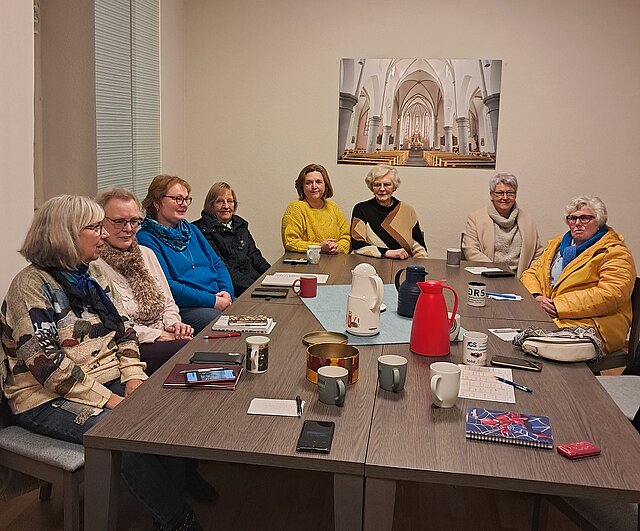 Gruppe von Frauen sitzt an einem rechteckigen Tisch in einem Raum mit einem großen Bild einer Kircheninnenansicht an der Wand