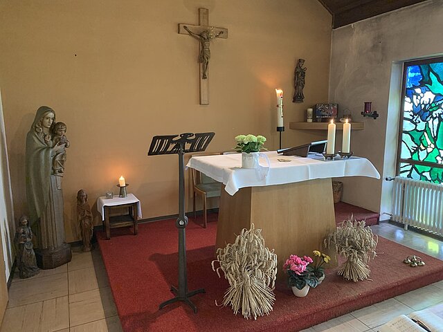 Altar mit weißem Tuch, Kerzen, Blumengestecken und Getreidebündeln in einer kleinen Kapelle mit Marienstatue, Kruzifix und buntem Glasfenster.