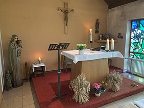 Altar mit weißem Tuch, Kerzen, Blumengestecken und Getreidebündeln in einer kleinen Kapelle mit Marienstatue, Kruzifix und buntem Glasfenster.