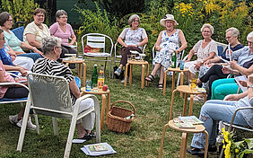 Eine Gruppe von Frauen sitzt auf Stühlen im Kreis an einem Sommertag in einem Garten, neben sich jeweils kleine Tische und darauf Trinkgläser und Getränkeflaschen.