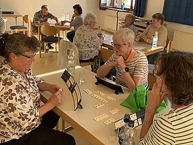 Mehrere ältere Personen sitzen an Tischen in einem hellen Raum und spielen Gesellschaftsspiele mit Spieltafeln und Karten