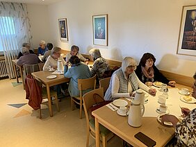 Mehrere Personen sitzen an zwei Tischen in einem hellen Raum und trinken Kaffee.