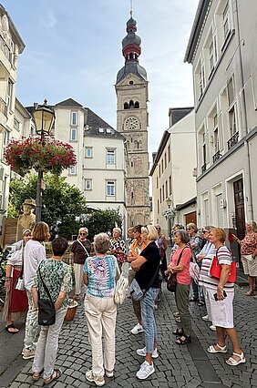 Gruppe von Menschen steht auf gepflastertem Platz vor einem hohen Kirchturm mit Uhr in einer Altstadt