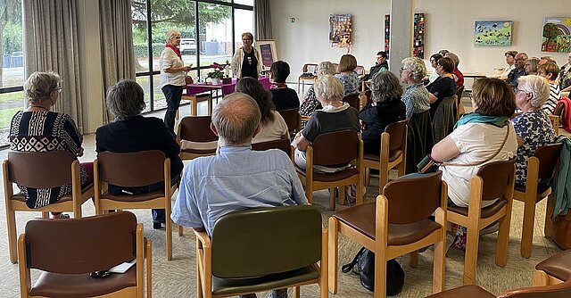 Eine Gruppe aus Frauen und Männern sitzt in einem Raum auf Stühlen, die im Halbkreis angebordneten sind. Vorne stehen zwei Frauen vor einem dekorierten Tisch und sprechen zur Gruppe.