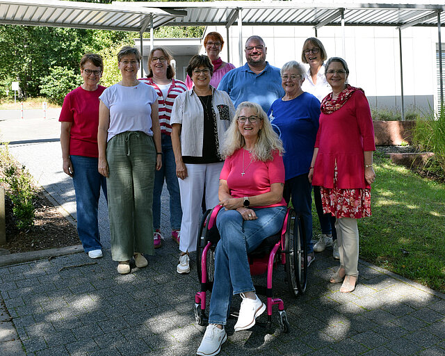 Gruppe von zehn Personen, neun stehen, eine Frau sitzt im Rollstuhl, alle lächeln, im Freien unter einem Vordach.