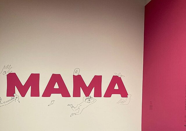 Foto des Eingangs zur Ausstellung mit dem Schriftzug "Mama" auf einer weißen Wand
