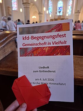 Liedheft mit der Überschrift 'kfd-Begegnungsfest Gemeinschaft in Vielfalt' und einem roten Papierherz in einer Kirche mit bunten Kirchenfenstern im Hintergrund.