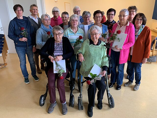 Gruppe älterer Frauen, zwei Frauen sitzen mit Rollatoren, mehrere halten rote Rosen und weiße Papiere, helle Innenraumumgebung
