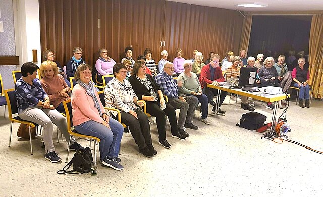 Eine Gruppe von Menschen sitzt in einem Saal. Die Stühle sind in drei Reihen hintereinander jeweils in einem Kreisbogen angeordnet. Die Gruppe ist gut gelaunt, einige Menschen schauen lächelnd in die Kamera.